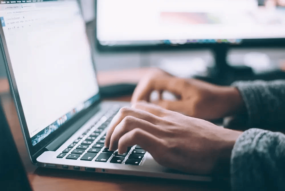Close-up of a person’s hands typing on a laptop keyboard, with two computer monitors in the background on a desk. The scene suggests working or studying in a modern, tech-focused environment.