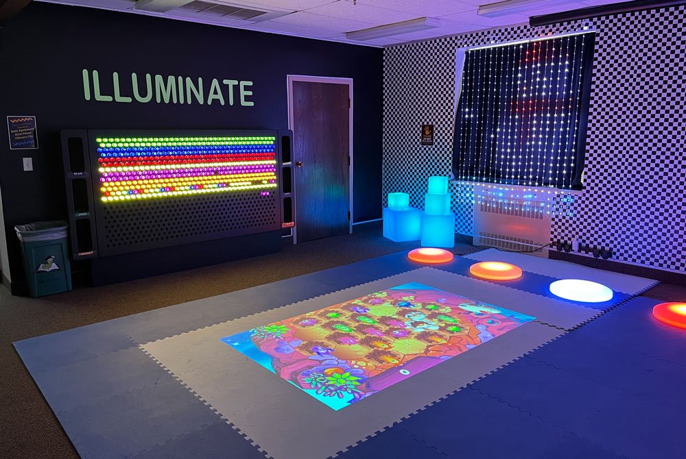 A sensory room with a large interactive light wall, glowing cube seats, a digital floor game, and the word "ILLUMINATE" on the wall, all lit with colorful LED lights.